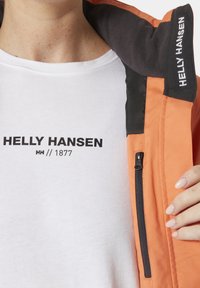 Oraange jas met zwarte accenten en een ritszak; gedragen over een witte T-shirt met een zwart logo en tekst. Zachte textuur zichtbaar.