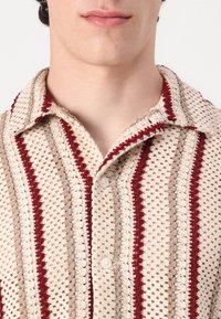 Cămașă cu mânecă scurtă în crem, cu dungi roșii și bej, având un model texturat din tricot și un guler tradițional cu bătăi.