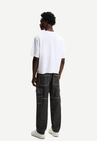 Witte T-shirt met korte mouwen gecombineerd met donkere cargobroek met meerdere zakken en contrasterende witte stiksels, tegen een effen achtergrond.