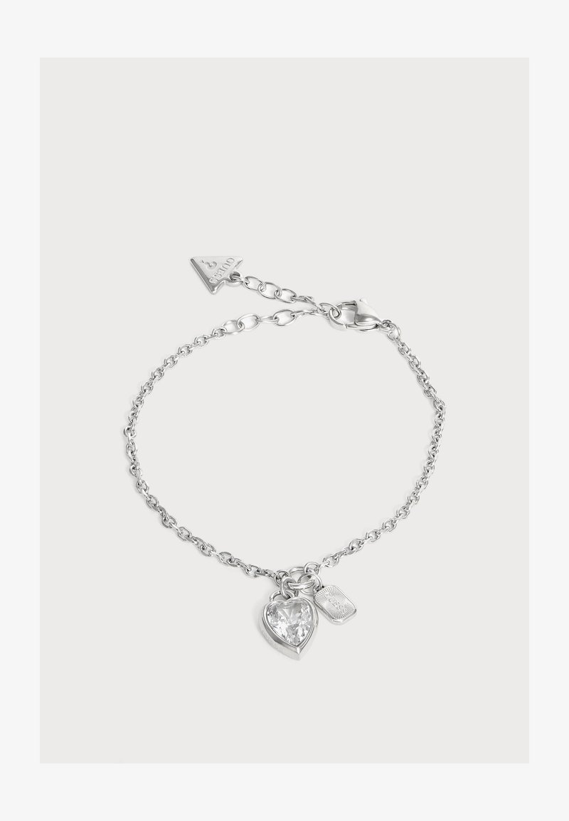 Bracelet chaîne en argent avec cristal clair en forme de cœur et petit charm rectangulaire, plus une étiquette logo Guess triangulaire près du fermoir.
