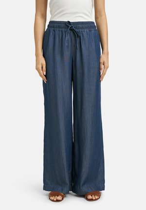 Frau trägt dunkelblaue, weit geschnittene Hose mit elastischem Bund und Kordelzug, kombiniert mit weißem ärmellosem Oberteil und braunen Sandalen mit offener Spitze.