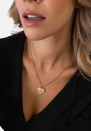 Frau trägt eine goldene Halskette mit einem herzförmigen Anhänger mit Augendesign, gekleidet in einem schwarzen V-Ausschnitt-Top.