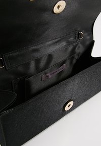Intérieur d'un sac en tissu noir avec une poche zippée et une étiquette portant la mention "ANNA FIELD". Le sac possède une fermeture à pression et une texture lisse.