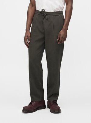Homme portant un pantalon vert olive foncé avec cordon de serrage et des bottes en cuir rouge foncé à lacets, debout devant un arrière-plan uni.