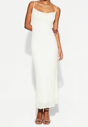 Kvinde iført en ærmeløs hvid blonde maxikjole og metalliske, snørede høje hæle, stående foran en ensfarvet baggrund.