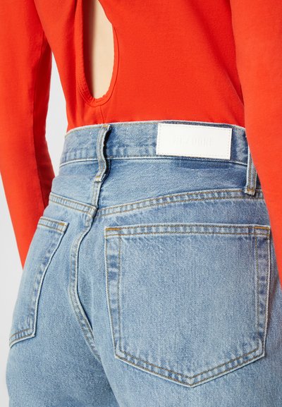 Top vermelho de mangas compridas com um recorte nas costas, combinado com jeans de cintura alta azul claro com um rótulo branco de marca e bolsos traseiros visíveis.