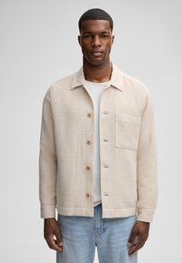 Giacca beige in tessuto con collo, chiusura a bottoni e taschino sul petto, abbinata a una camicia bianca e jeans azzurri chiari.