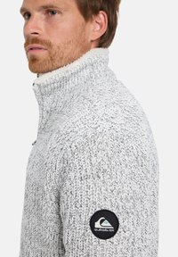 Hellegrau gestrickter Pullover mit hohem Kragen, ausgestattet mit einem weichen Fleecefutter und einem schwarzen runden Logopatch auf dem linken Ärmel.