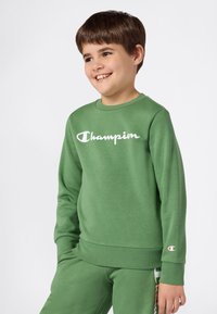 Jongeman in een groene Champion sweatshirt en bijpassende broek, glimlachend met handen in zijn zakken, voor een effen witte achtergrond.