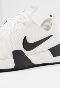 Valkoinen urheilukenkä, jossa on verkkopinta, mustalla swoosh-logolla ja teksturoidulla mustalla kantapäällä. Tasainen valkoinen pohja.