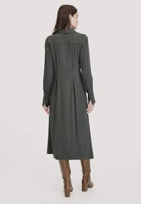 Eine Frau trägt ein dunkelgrünes, langärmeliges Midikleid und braune knielange Stiefel. Sie steht und schaut von der einfachen Hintergrund weg.