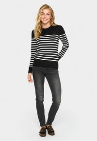 Maglione a righe bianche e nere con collo rotondo e polsini a coste, abbinato a jeans scuri e scarpe marroni. Tessuto morbido, design semplice.