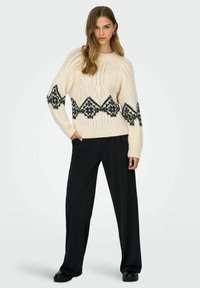 Crèmekleurige gebreide trui met een zwart geometrisch patroon rond het midden en de mouwen. Gecombineerd met loszittende zwarte broek.
