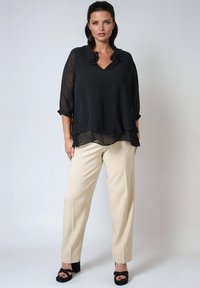 Zwarte, gelaagde blouse met ruches bij de hals, driekwart mouwen; gecombineerd met beige, losse broek en zwarte hoge hakken.