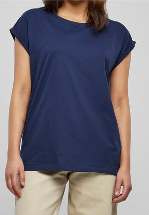 Person trägt ein einfaches marineblaues T-Shirt mit kurzen Ärmeln und aufgerollten Bündchen sowie helle beige Hosen und steht vor einem einfarbigen Hintergrund.