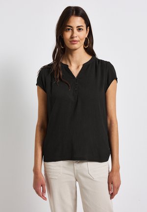 Blusa nera a maniche corte con scollo a V, realizzata in tessuto strutturato e con tre bottoni sul davanti. Indossata con pantaloni di colore chiaro.