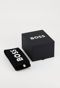 Boîte noire avec le logo "BOSS" en blanc sur le dessus, accompagnée d'une pochette noire avec "BOSS" imprimé en blanc, présentant un détail doré et rayé.