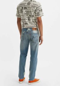 Jeans de mezclilla azul deslavado con corte de pierna recta, combinados con una camisa de manga corta estampada y zapatillas naranjas. Detalle de parche de cuero marrón.