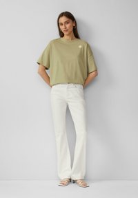 Olijf groene oversized t-shirt met een kleine zon graphic, gecombineerd met witte flared jeans en witte sandaaltjes met bandjes. Gladde stoftextuur.
