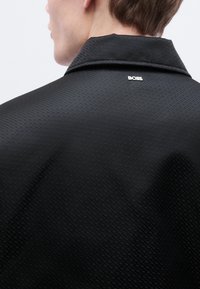 Μαύρο υφασμάτινο μπουφάν με διακριτικό γεωμετρικό σχέδιο. Διαθέτει μυτερό γιακά και ασημένιο λογότυπο "BOSS" κοντά στον λαιμό.