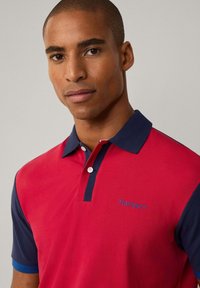 Camisa polo roja y navy con cuello, dos botones y un logo bordado en azul en el lado izquierdo delantero. Textura suave.