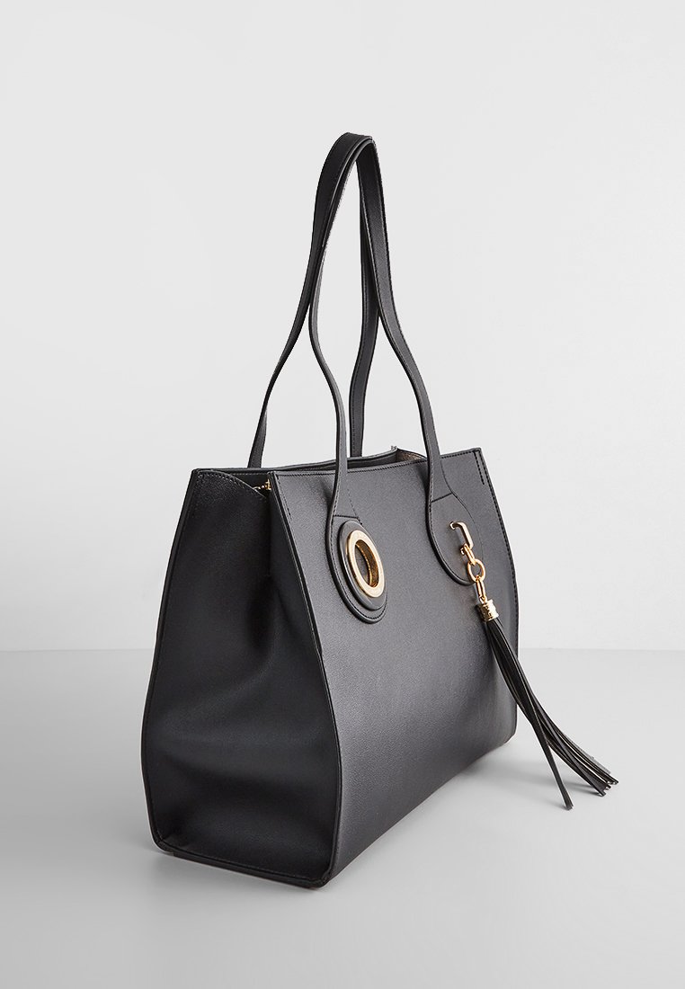 Sac cabas en cuir noir avec une forme structurée, mettant en avant un accent en métal doré en forme circulaire et un détail de pompon sur la tirette de la fermeture éclair.