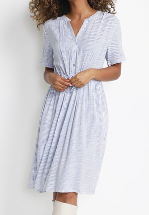 Frau in einem knielangen, kurzärmeligen, blau-weiß gestreiften Kleid, die die Taillenschleife vor einem schlichten weißen Hintergrund justiert.