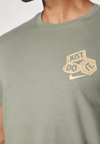 Camiseta de algodón verde oliva de manga corta, con un escote redondeado y un gráfico del logo "Just Do It" en color beige en el pecho.