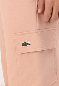 Lyserøde cargo-bukser har et tekstureret stof, sidelomme med klap og broderet grøn krokodille-logo ved lommens kant.
