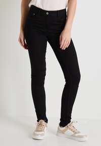 Jeans skinny negros hechos de un material elástico, con un diseño de cintura alta, cinco bolsillos y cierre de botón en la parte delantera, combinados con zapatillas ligeras.