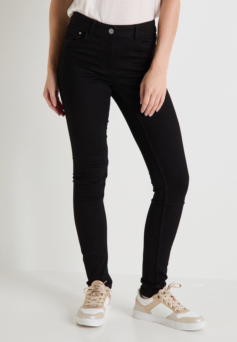 Jeans skinny negros hechos de un material elástico, con un diseño de cintura alta, cinco bolsillos y cierre de botón en la parte delantera, combinados con zapatillas ligeras.