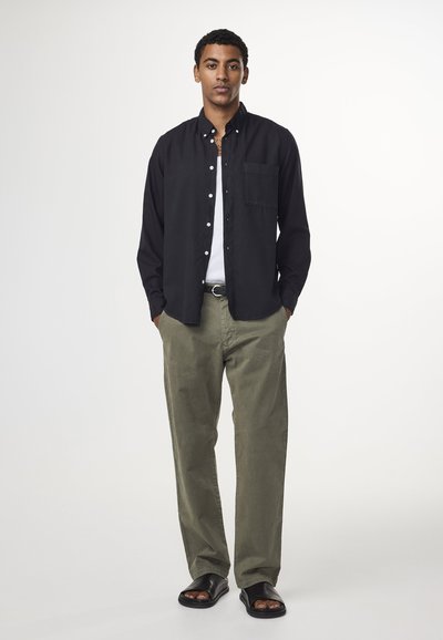 Chemise noire à boutons, T-shirt blanc en dessous, pantalon vert olive et sandales noires. Les détails clés incluent des boutons, une poche poitrine et une coupe ample.