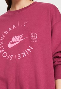 Nike sweatshirt i en djup rosa färg, med vit grafisk text och logotyp. Designen inkluderar cirkulära textelement och en rundad halsringning.