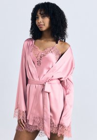 Γυναίκα με σγουρά μαλλιά που φοράει ρόμπα από ροζ σατέν και δαντέλα και φόρεμα-εσώρουχο, όρθια μπροστά σε απλό ανοιχτόφωτο φόντο.