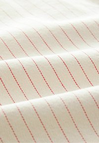 Tejido de algodón con un fondo crema y finas rayas verticales rojas. La textura parece suave y lisa, mostrando un patrón tejido.