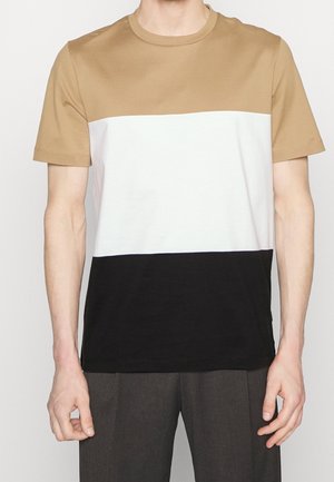 Man draagt een T-shirt met korte mouwen en ronde hals met horizontale kleurvlakken in beige, wit en zwart, gecombineerd met een donkere broek.