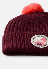 Béret en tricot bordeaux avec une texture côtelée, surmonté d'un pompon rose duveteux. Présente un patch circulaire avec des graphismes de montagne.