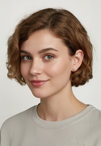 Silverfärgade stjärnformade studs, texturerat material, medelhög glans, bärs av en modell med lockigt hår som har på sig en ljusgrå skjorta.