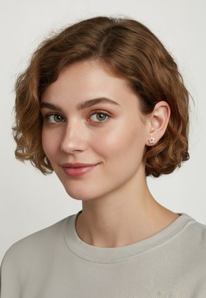 Silverfärgade stjärnformade studs, texturerat material, medelhög glans, bärs av en modell med lockigt hår som har på sig en ljusgrå skjorta.