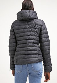 Svart pufferjacka med horisontellt sydda paneler och huva. Texturerat material, åtsittande design, kombinerad med blå jeans.