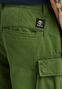 Zelené cargo šortky s jemnou texturou, několika kapsami a nášivkou Timberland na zadní straně. Prošité detaily v odpovídající nitě.