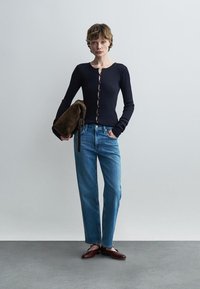 Γυναίκα που φοράει μαύρο μακρυμάνικο τοπ με κουμπιά, μπλε ψηλόμεσα τζιν, μπορντό φλατ παπούτσια, κρατώντας καφέ τσάντα από σουέτ, στέκεται μπροστά σε απλο γκρι φόντο.