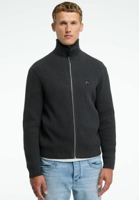 Schwarzer gerippter Strick-Zipper mit hohem Kragen, ausgestattet mit einem dezenten Logo-Patch auf der Brust und einer strukturierten Oberfläche.
