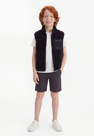 Rødhåret dreng iført en sort zip-up vest over en hvid t-shirt, mørke shorts og hvide sneakers, smilende med den ene hånd i lommen.