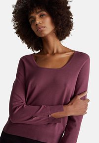 Frau mit lockigem Haar, die einen langärmeligen violetten Pullover mit quadratischem Ausschnitt trägt und einen Arm vor ihrem Körper hält.