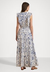 Maxime-mekko sinisellä kukkakuviolla beige-kankaalla, röyhelöhihoilla, joustovyötäröllä ja kerroksellisella muotoilulla. Jalkineena on litteäpohjaiset sandaalit.