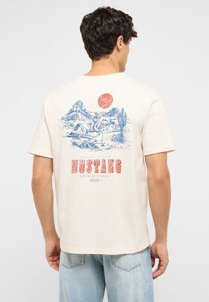 Man wearing cream t-shirt with blue and red western landscape print featuring rider on horse, cactus, mountains, and sun, paired with light blue jeans.