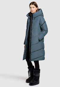 Kvindelig iført en lang teal pufferjakke med hætte, sorte tights og sorte vinterstøvler, stående mod en ensfarvet grå baggrund.