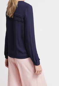 Blusa a maniche lunghe blu navy con dettagli a pieghe sul retro, realizzata in tessuto morbido e con polsini a bottoni, abbinata a pantaloni larghi rosa pallido.