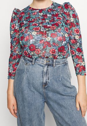 Blomstret bluse med lange, gennemsigtige ærmer, der har en blanding af røde, blå og grønne blomster mod en gennemsigtig baggrund. Højtaljede lyse denim jeans.